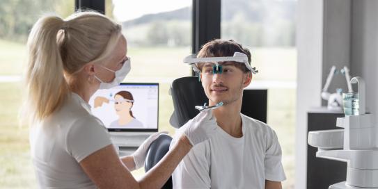 Zahnärztin justiert ein Gesichtsbogen-Messgerät bei einem jungen Patienten in moderner Praxisumgebung.