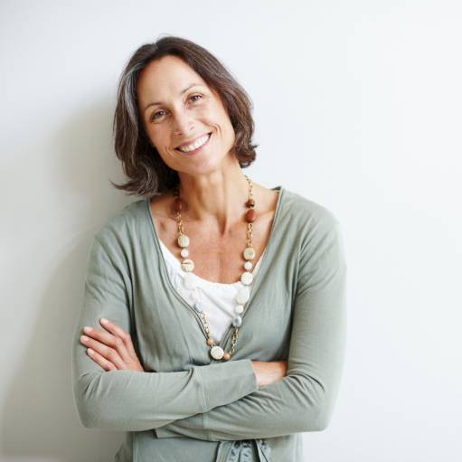 Reifere Frau mit halblangen braunen Haaren steht lächelnd mit verschränkten Armen vor einer weißen Wand, trägt eine lange Kette und einen hellgrünen Cardigan.