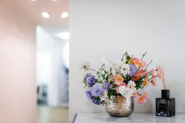 Bunter Blumenstrauß in einer strukturierten silbernen Vase auf einem weißen Sideboard, daneben ein Raumduftspender in einem dunklen Glasflakon.