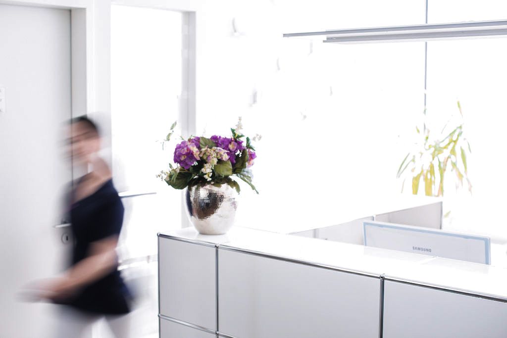 Heller Empfangsbereich einer Zahnarztpraxis mit Blumendekoration auf dem Tresen und unscharfer Bewegung einer vorbeigehenden Person.