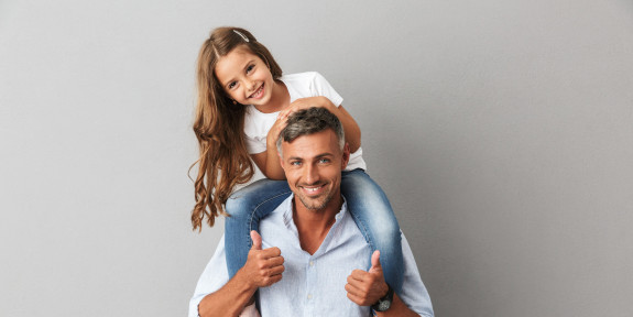 Glücklicher Vater mit seiner Tochter auf den Schultern, beide lächeln in die Kamera, vor grauem Hintergrund.